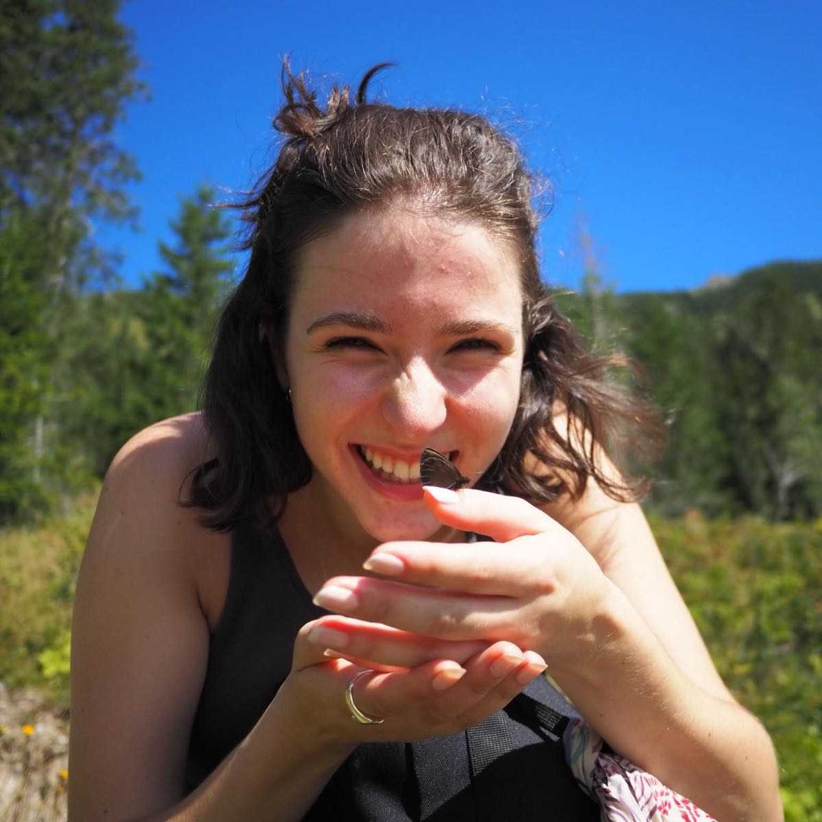 Photo of Weronika Korpak. Smiling woman holding a butterfly with a clear blue sky and greenery in the background.