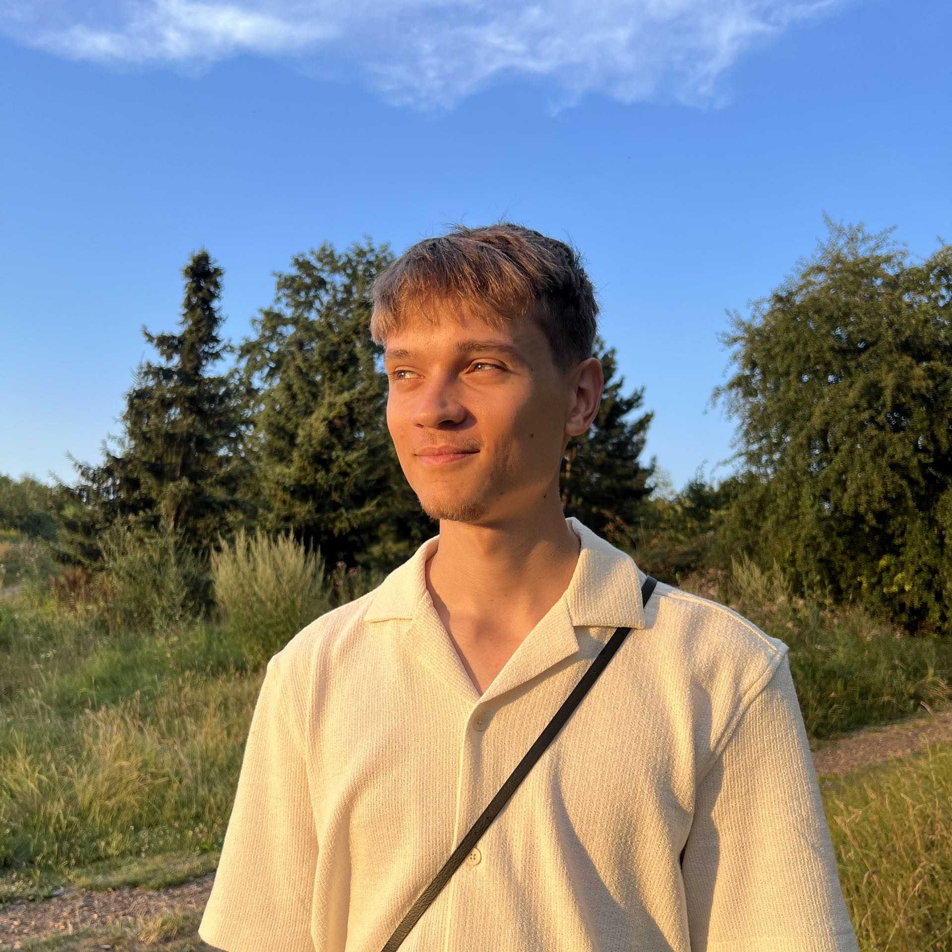Photo of Michał Zuch. Young man standing outdoors, bathed in warm sunset light, with trees and a clear sky in the background.