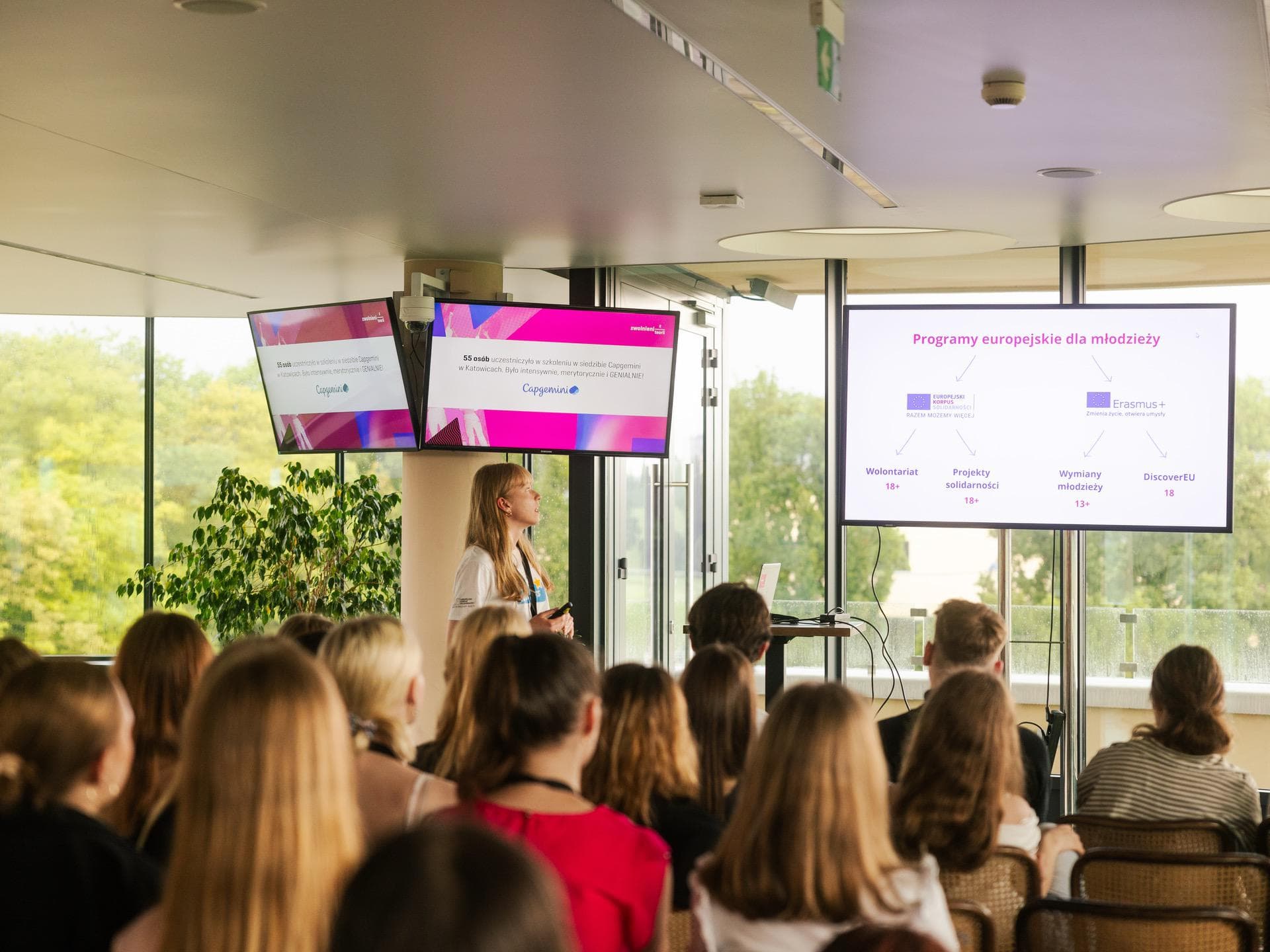 Prelegentka prezentująca przed publicznością w nowoczesnym wnętrzu. Dwa ekrany wyświetlają informacje o europejskich programach dla młodzieży. Publiczność, głównie młodzi ludzie, uważnie słucha. W tle duże okna pokazują zieleń na zewnątrz.