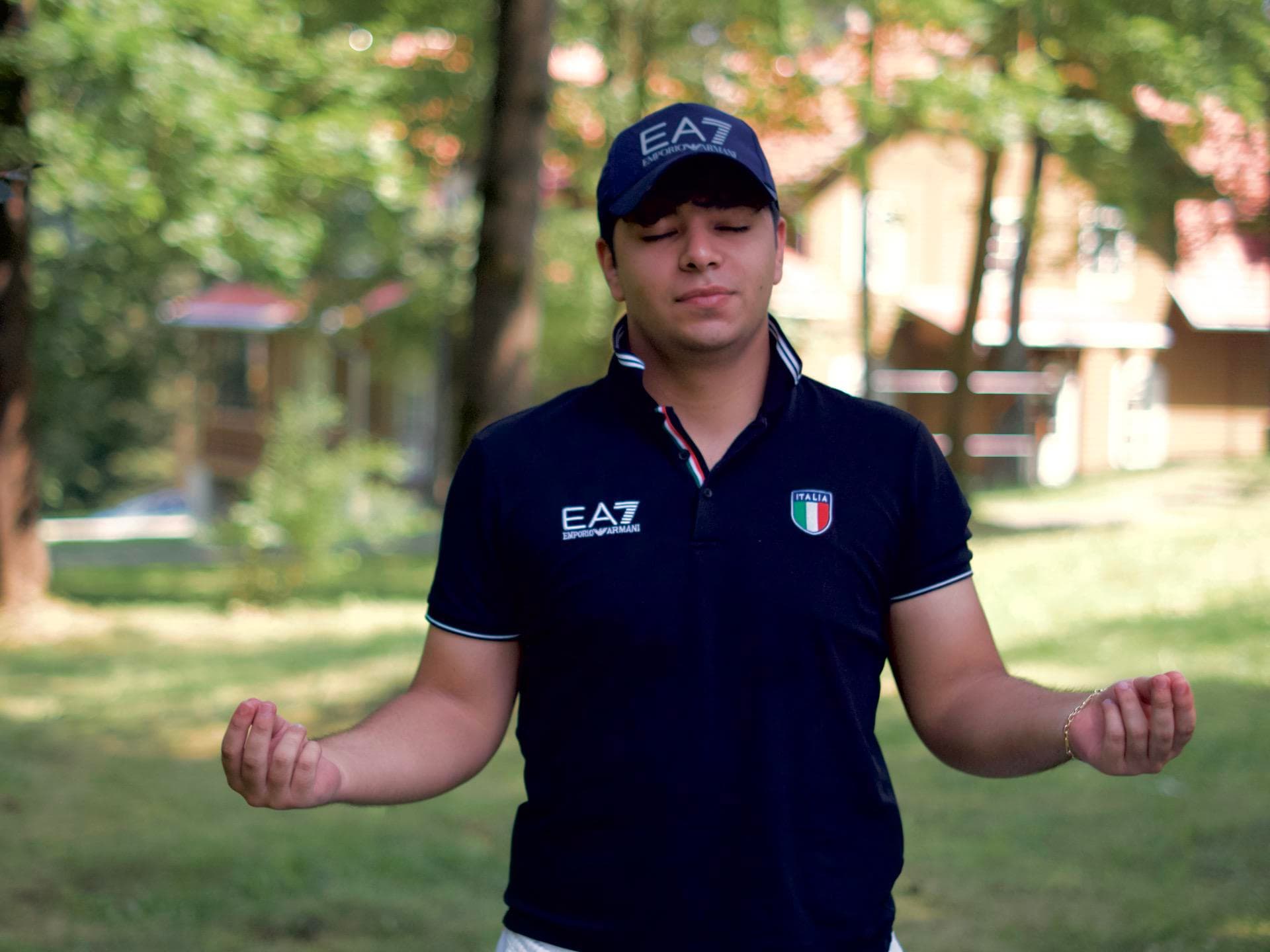 Młody mężczyzna stojący na świeżym powietrzu z spokojnym wyrazem twarzy, zamkniętymi oczami i rękami w pozycji medytacyjnej. Ma na sobie ciemną koszulkę polo i dopasowaną czapkę. W tle widać rozmyte drzewa i budynki, co sugeruje spokojne otoczenie naturalne.